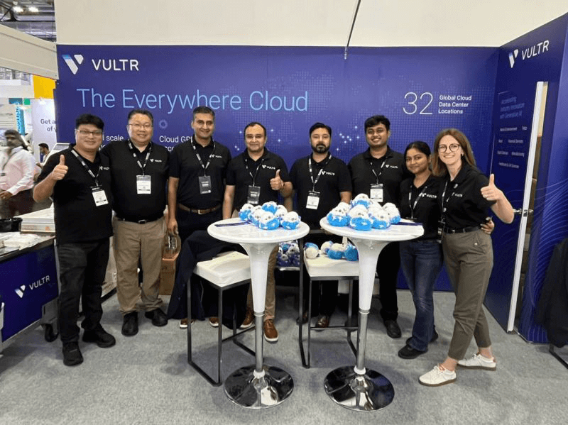 A team of Vultr employees standing at their exhibition booth at a tech conference. They're wearing matching black polo shirts with Vultr logos, giving thumbs up, and posing behind display tables with Morty, the Vultr mascot, plushies. The backdrop features the Vultr logo and 'The Everywhere Cloud' tagline.