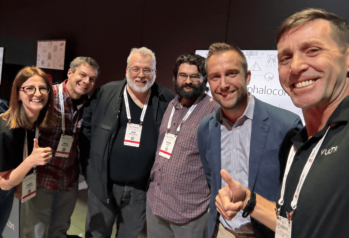 Six Vultr employees standing in a row at CephaloCon, all smiling at the camera. They're wearing conference badges on lanyards. A white CephaloCon backdrop with simple line drawings is visible behind them. The woman on the far left is wearing glasses and giving a thumbs up, while the man on the far right has a Vultr logo visible on his black polo shirt. The group represents a diverse mix of ages.