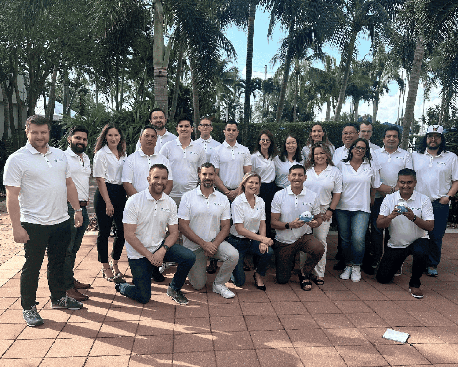A large group photo of Vultr team members with palm trees in the background. Everyone is wearing matching white polo shirts with the Vultr logo. The team is arranged in two rows on a brick paver patio, with some standing and others kneeling in the front row.