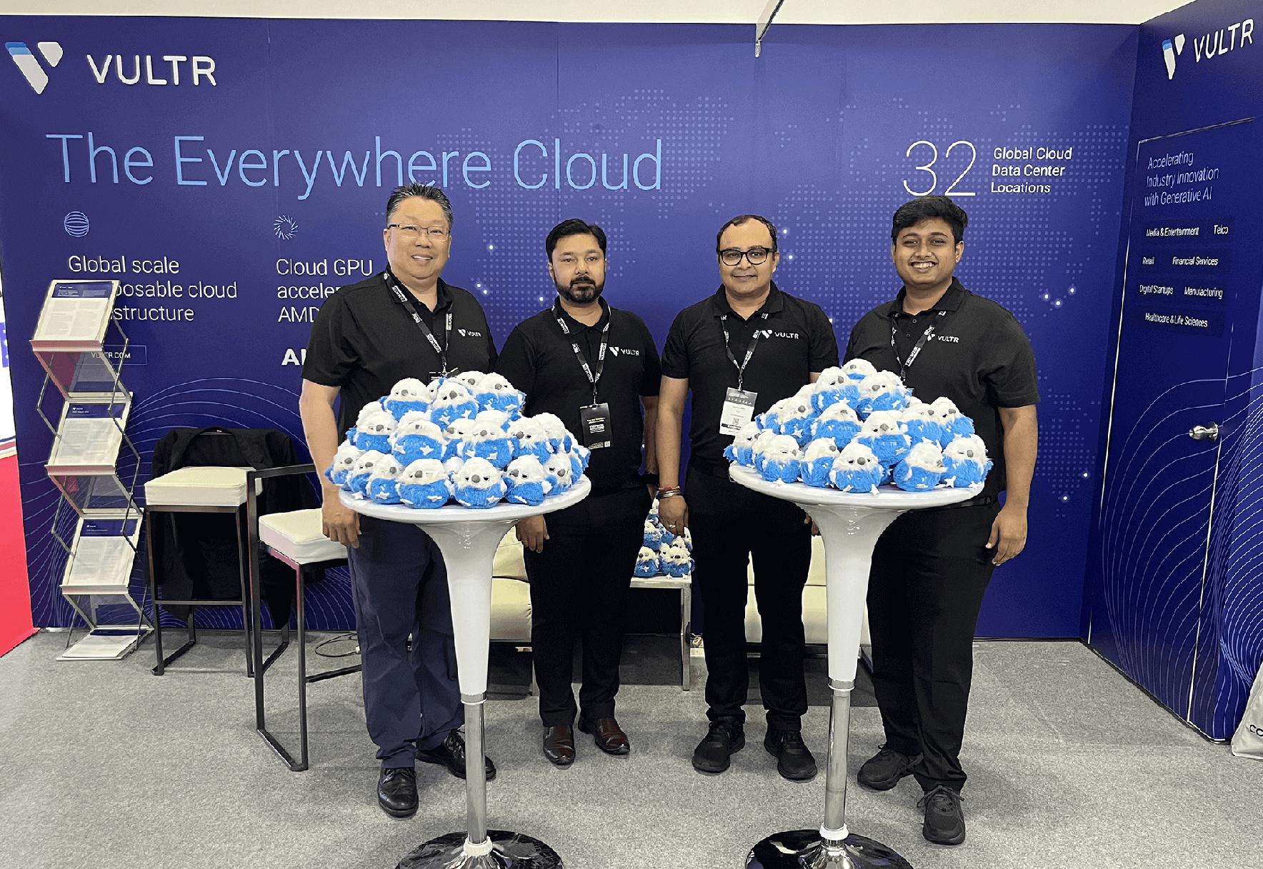 Four Vultr employees standing at their exhibition booth, wearing matching black polo shirts with Vultr logos. They're positioned between two white pedestal tables displaying numerous small blue and white Morty plushies. Behind them is the company's blue backdrop featuring 'The Everywhere Cloud' slogan, mentions of '32 Global Cloud Data Center Locations,' and a dotted world map design.