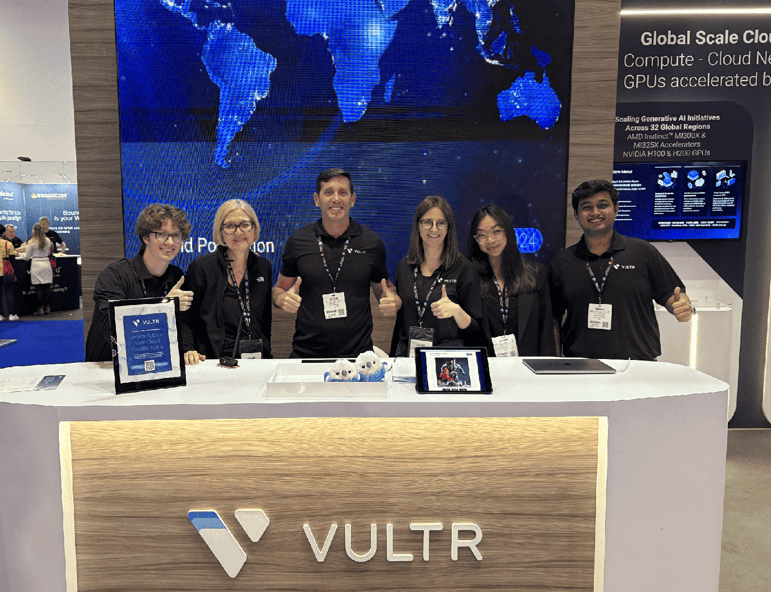 Vultr booth staff at a tech conference exhibition. Six people wearing black Vultr-branded shirts are standing behind a wooden counter with the Vultr logo, all giving thumbs up. Behind them is a large digital display showing a blue world map and text about 'Global Scale Cloud' with information about GPU accelerators and AI initiatives across 32 global regions.